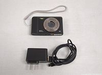 Digital camera with lens, wrist strap, USB charging cable, and power adapter visible.