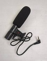 View of Digipower microphone from one side showing foam windshield, mounting bracket, and cable with 3.5mm jack.