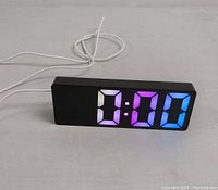Front view of black digital LED clock showing 0:00 with multicolor digits on a white surface with power cable attached.