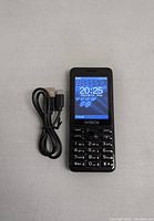 Front view of the black keypad mobile phone powered on displaying time and date, with USB charging cable coiled on left side