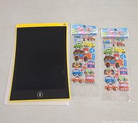 Full view of the yellow-framed LCD writing tablet alongside two identical packets of vehicle-themed stickers showing cars, trucks, buses, motorcycles, and traffic signs