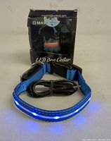 Blue LED dog collar lit and displayed with USB charging cable in front of original box with branding and LED dog collar text.