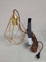 Side angle view of one gold-plated geometric pendant light with twisted fabric cord and black mount lying on floor against wall.