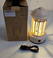 Mini lamp lit emitting a warm light, standing beside its original box and a USB cable for charging. The lamp is beige with vertical slats around the light source and a power button on its base.