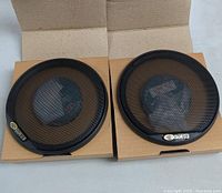 Front view of two SAM US 4-way speakers with black metal mesh grills in cardboard boxes, showing speaker grills and SAM logo.