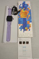 Purple smart watch in silicone strap with black screen in box, charging cable, and manual.