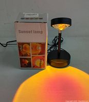 Sunset lamp turned on showing warm orange and pink light projection on surface with original box showing product images.