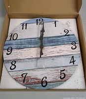 Front view of wall clock in box showing blue, white, and brown striped wooden plank design with black numbers and black clock hands.