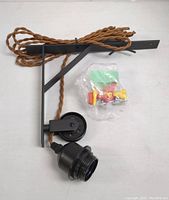 Black wall mount fixture with twisted brown cord attached and mounting hardware bag included, displayed on white surface.