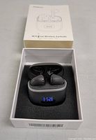 Photo showing open box with black wireless earbuds inside charging case. Charging case displays battery percentage on LED screen. Earbuds have silicone tips.