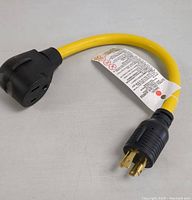 Yellow generator adapter cable with black 4-prong locking male plug and black 4-prong locking female connector end with attached safety tag, laying on white surface.