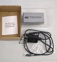Photo of 4K HD Audio Extractor device, power adapter, cable, and user manual laid out next to open cardboard box.