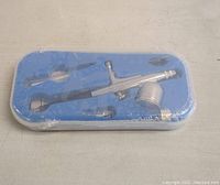 Airbrush spray tool kit components arranged inside a blue molded plastic tray covered in plastic wrap, showing metallic airbrush gun and accessories