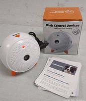 White circular bark control device with orange button, red indicator light, perforated front, USB cable, troubleshooting guide, and original box showing product image.