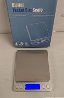 Photo of the digital pocket size scale next to its original blue box packaging with product image and text.