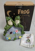 Photo of the front of the Frog Solar Statue showing two green frogs sitting on a rock with colorful flowers and 'WELCOME' text, with original box behind.