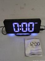 Front view of LED digital alarm clock showing blue LED time display 0:00 with day (MON). Clock is black rectangular with a curved base, placed on white surface next to folded instruction manual.