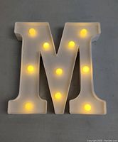 Letter M standee illuminated with warm yellow LED lights on a flat grey surface.