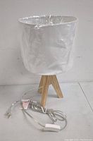 Table lamp with light wood tripod base and white fabric cylindrical shade, wrapped shade plastic still on. Power cord with inline switch visible.