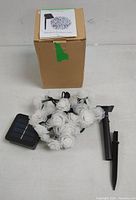 Photo of the solar string lights showing white rose-shaped LED covers, solar panel, and stake, with the packaging box.