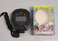 Photograph of the black XL-009B digital stopwatch alongside its original colorful box displaying runners at a starting line, highlighting the device's compact size and design.