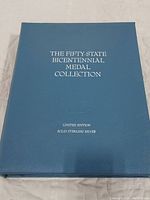 Front cover of blue hardcover binder with silver foil title “The Fifty-State Bicentennial Medal Collection” and “Limited Edition Solid Sterling Silver.”