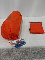 Orange cylindrical storage bag with Big Agnes logo containing the tent and a smaller orange pouch for the footprint ground cloth on a white quilt.