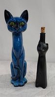 Full view of tall blue ceramic cat figurine beside black cat glass bottle decanter with cork stopper