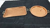 Two copper serving trays laid side by side on a black fabric background, one rectangular and one round, showing their shape and handles.