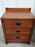 Front view of the nightstand showing three drawers with black metal drop pull handles and solid wood construction. Small discoloration mark on the top left side of surface visible.