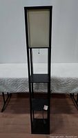 Full view of black wood floor lamp with shelves and off white shade, showing overall shape and shelving.