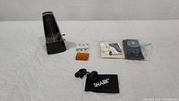 Mechanical metronome, Snark clip-on guitar tuner pouch, electronic tuning devices and other guitar accessories laid out on white surface.