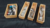 Four wooden trays displaying sets of stainless steel knives, forks, spoons, and one tray with mixed metal and plastic kitchen utensils including strainer, corkscrew, can opener, and measuring spoons.