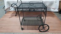Full side view of the black wrought iron tea cart showing two shelves, handle, wheels and caster wheels.