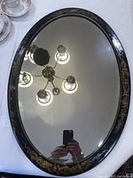 Full view of oval mirror showing glass and black lacquered wooden frame with gold leaf floral decorations reflecting ceiling light fixture.