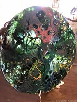 Top angled view of round art glass plate showing multicolored fused glass texture with green, red, yellow and brown bubbly abstract patterns and glossy finish.