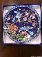 Top view of the cloisonné plate showing birds and flowers design on a dark blue background inside a box.