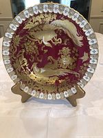 Front view of a decorative plate with gold birds and floral pattern on maroon background, displayed on a stand