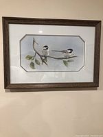 Framed watercolor painting showing two black-capped chickadee birds on a branch with leaves, framed in a dark wood frame against a light wall.