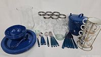 Photo showing entire kitchenware set including blue enamel stockpot with lid, ceramic dishes, flatware, glass goblets, vase, jar, and stacking mugs with rack.
