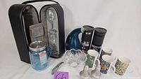 Full view of entire kitchenware and drinkware assortment including travel mugs, Mason tumbler, wine glasses in case, oil jug, salt and pepper shakers, glass bowl, and colorful ceramic mugs.