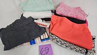Overview of 10 clothing items, folded, showing variety of patterns and colors including pink leaf-patterned shorts, coral pants, grey pants, and pastel tops.