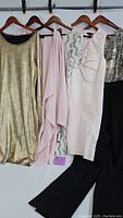Four women's dresses hanging on wooden hangers, showing color and styles: metallic gold, light pink chiffon, light pink with sequins, and black with metallic top.