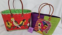 Two large embroidered tote bags displayed side by side. One has bird and floral embroidery on a red background; the other features an abstract face embroidery on a purple and green background. Both have brown leather handles.