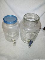 Two clear glass drink dispensers shown front view, one with a blue lid and white spigot, the other with textured surface and blue spigot.