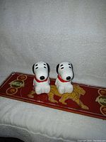 Photo showing pair of Snoopy salt and pepper shakers sitting facing forward on a red MGM tray.