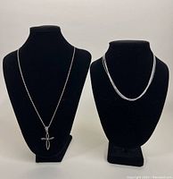 Front view of two necklaces displayed on black stands. Left is a silver chain with a cross pendant, right is a four-strand silver necklace.