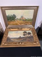 Two framed oil paintings stacked, one with a silver textured frame and countryside rural scene, the other in ornate gold frame with moody landscape