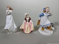 Front view of three porcelain figurines: a woman in white dress, a girl in pastel dress with bonnet, and a girl in blue dress with dog and satchel