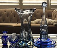 Full frontal display of all glassware showing decanter with stopper, clear and blue crystal vase, cobalt blue bud vase, and blue trivets on glass table.